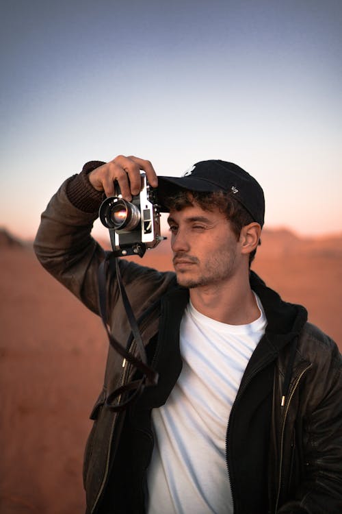 Focused young male traveler in stylish outfit and cap taking photo on vintage camera during trip in desert valley during sunset