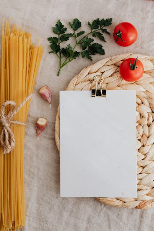 From above of blank paper with clip on wicker table mat among raw spaghetti and fresh tomatoes with parsley and cut garlic on linen surface