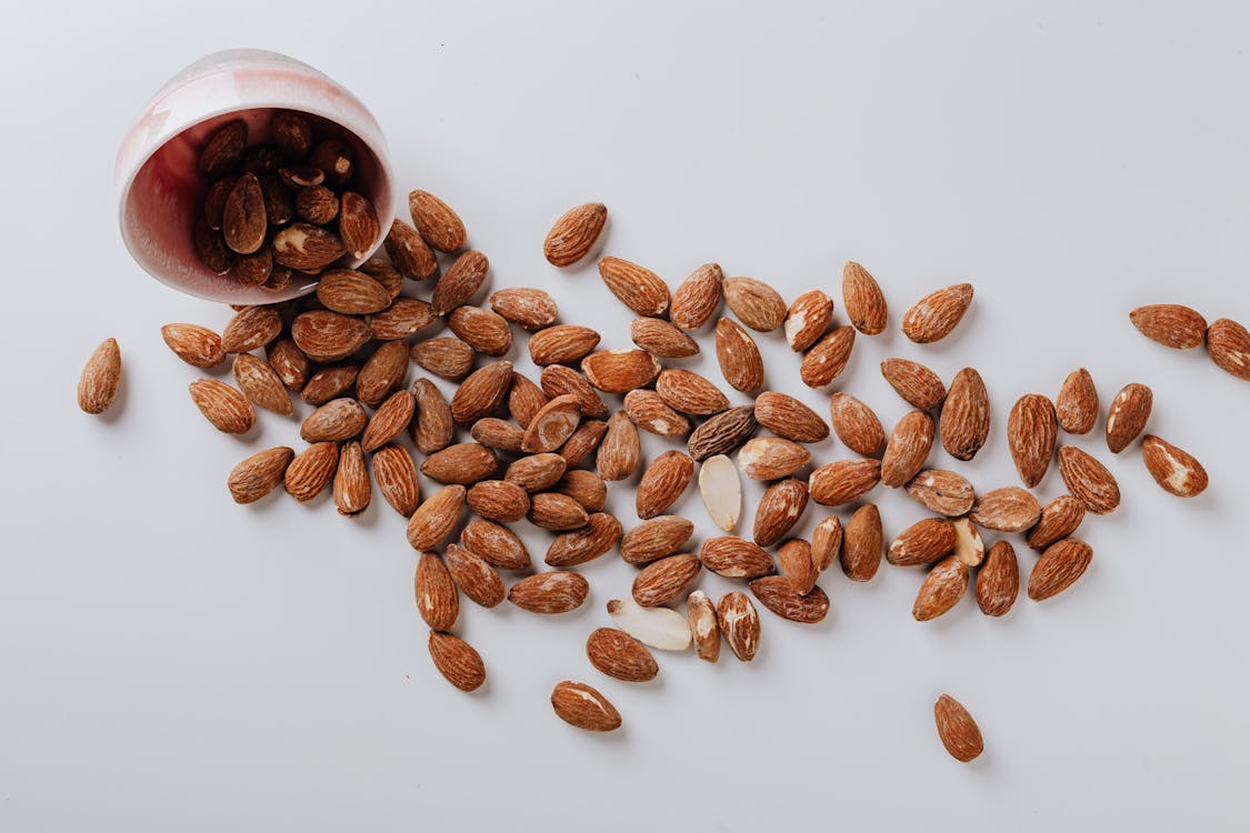 From above of raw oval shaped brown almonds scattered from small deep ceramic bowl on white surface