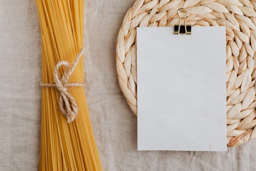 Free From above of blank paper placed on wicker table mat next to spaghetti tied with bow arranged on table covered with linen tablecloth suitable for recipe or ingredient listing Stock Photo