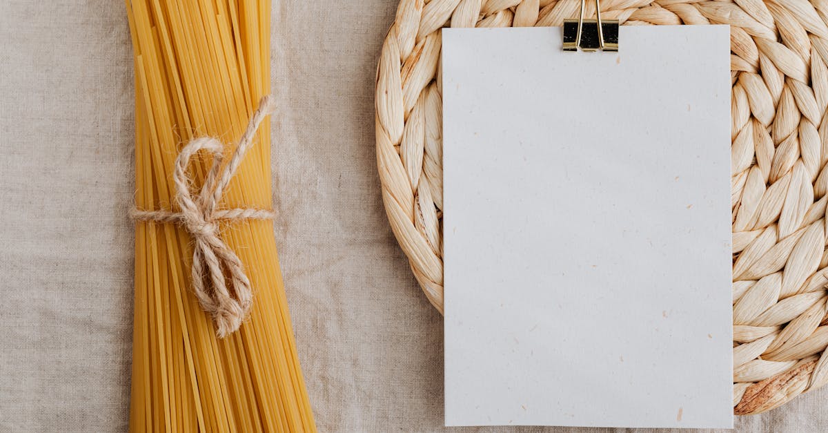 From above of blank paper placed on wicker table mat next to spaghetti tied with bow arranged on table covered with linen tablecloth suitable for recipe or ingredient listing