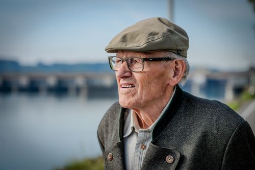 Free Senior man wearing a hat and glasses looks thoughtfully at a river scene in Wallsee, Austria. Stock Photo