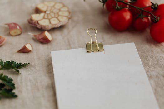 From above of empty clipboard for recipe ingredients or menu placement located on desk with linen tablecloth and surrounded by fresh delicious tomato on branch together with chopped garlic and green parsley