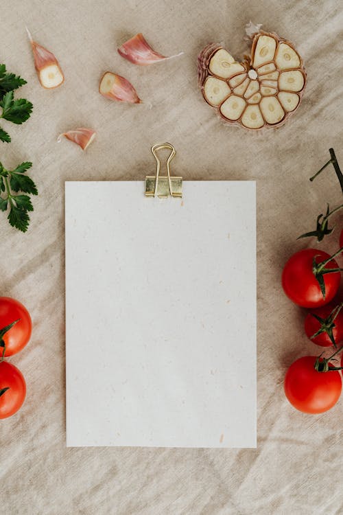 Free Top view of empty clipboard with golden paper binder placed on linen tablecloth among delicious red tomatoes on branches together with chopped garlic and green parsley suitable for recipe or menu placement Stock Photo