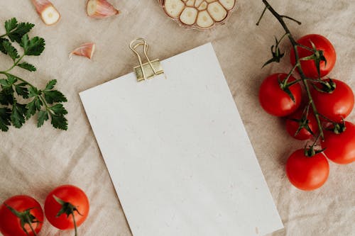 Imagen de un portapapeles en blanco con un sujetapapeles de papel dorado colocado sobre un mantel de mesa de lino entre tomates rojos deliciosos en ramas junto con ajos cortados y perejil verde dedicado para colocar recetas o menús