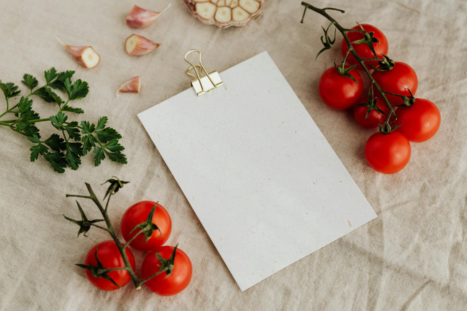 Top view of blank clipboard with golden paper binder placed on linen tablecloth among tasty red tomatoes on branches together with chopped garlic head and green parsley ideal for recipe or menu
