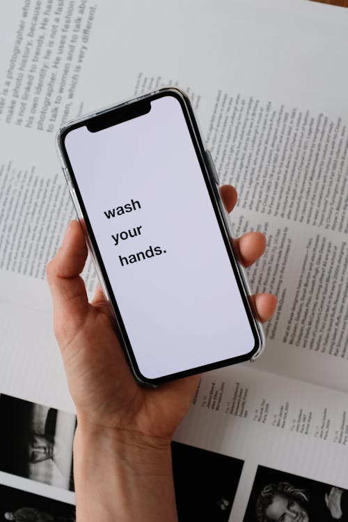 Free From above of unrecognizable person taking modern cellphone with WASH YOUR HANDS inscription on blank screen placed under page of book illustrating hygiene concept Stock Photo