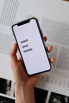 From above of unrecognizable person taking modern cellphone with WASH YOUR HANDS inscription on blank screen placed under page of book illustrating hygiene concept