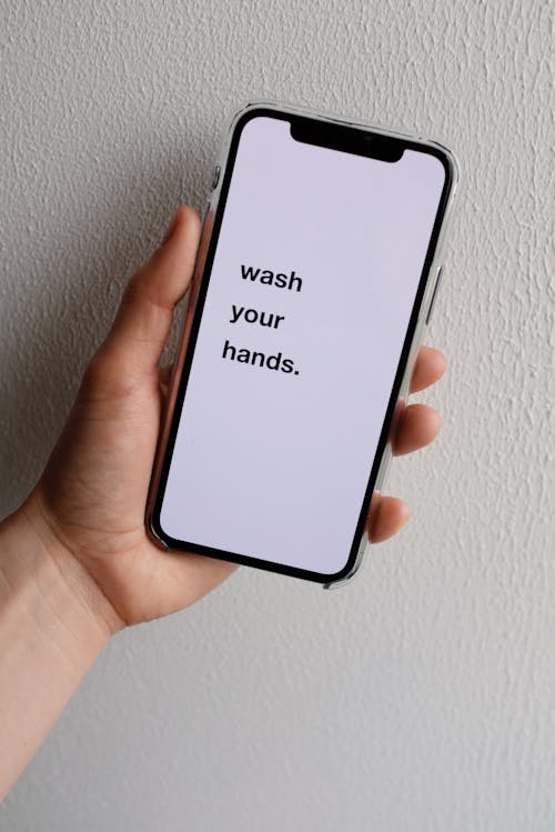 Crop anonymous person demonstrating modern cellphone screen with WASH YOUR HANDS inscription on white background with uneven surface in daylight during COVID 19 pandemic