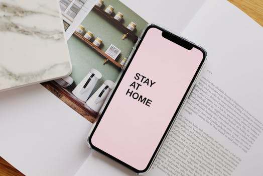 From above of modern cellphone with pale pink screen and STAY AT HOME inscription put on open magazine in apartment during COVID 19 pandemic