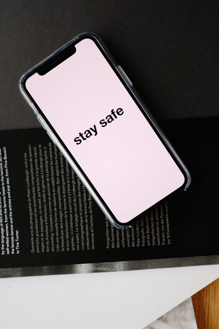 Top view of modern mobile phone with pale pink screen and STAY SAFE inscription placed on open magazine with black page and white letters during COVID 19 pandemic