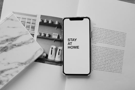Monochrome flat lay featuring a smartphone with 'Stay at home' message on an open book, promoting home learning.