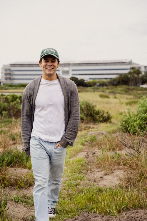 Free Young man in casual attire walking through grassy terrain with a modern building in the background. Stock Photo