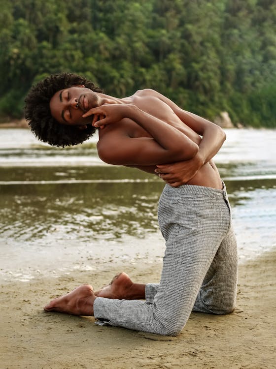 Topless Man in Gray Pants Kneeling on Sand Near Body of Water