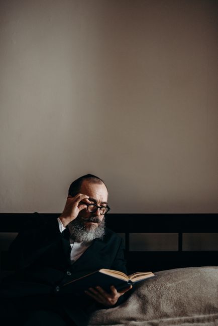 A bearded Orthodox Jewish man reading a book in a dimly lit room, reflecting on tradition.