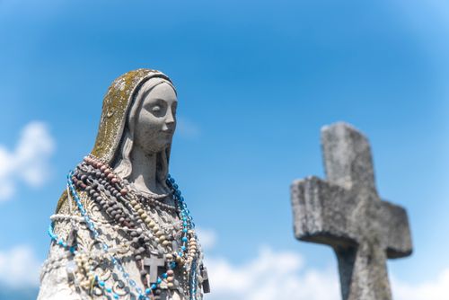 Free Statue of a religious figure adorned with rosary beads beside a stone cross and clear blue sky. Stock Photo