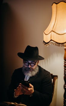 Elderly man in hat using smartphone, sitting by vintage lamp indoors.
