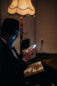 Orthodox Jewish man in hat reading book and using smartphone under a warm-lit lamp indoors.