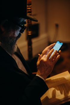 Orthodox Jewish man reading a book while using a smartphone indoors.