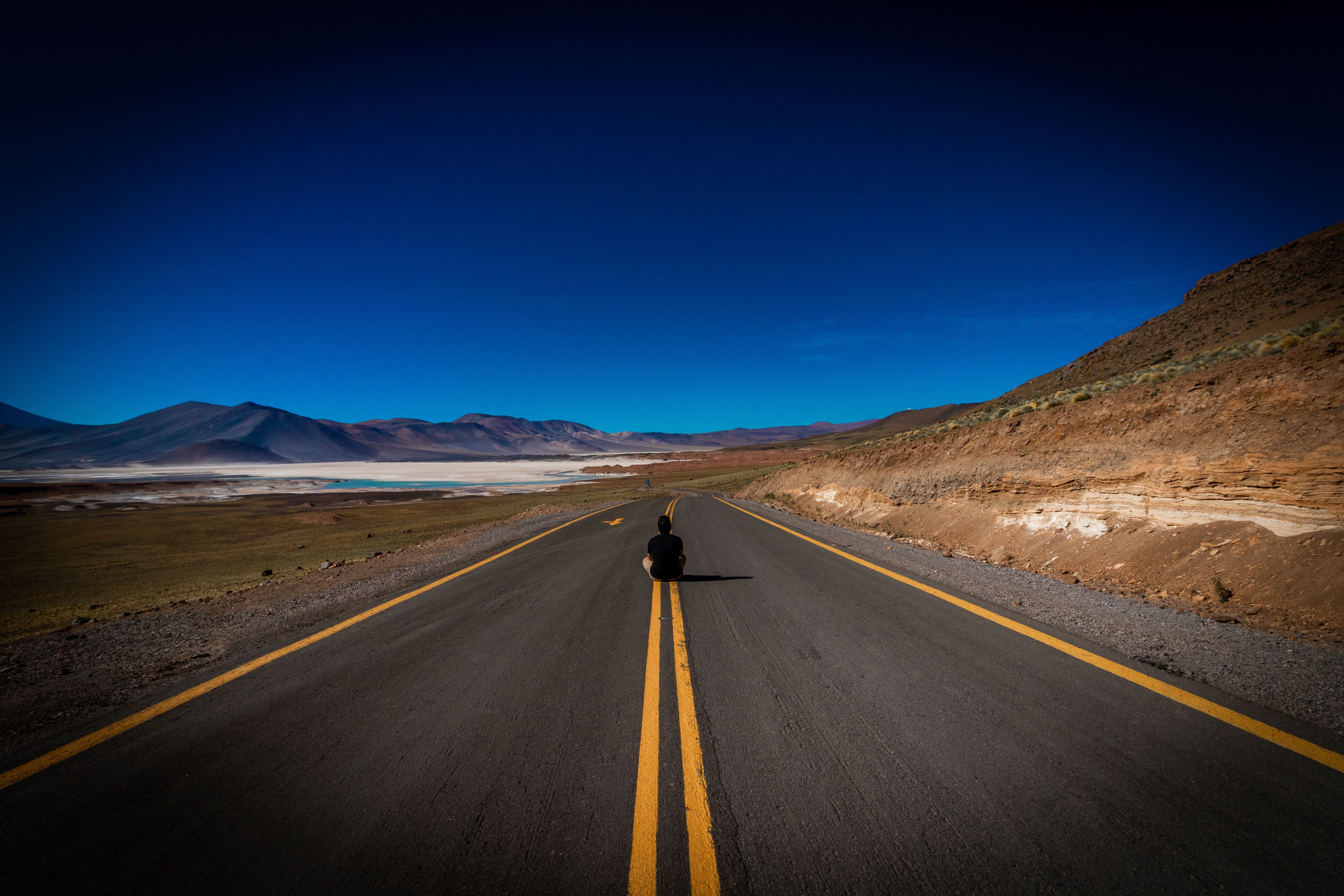Lonely traveler sitting on a vast desert highway under a clear blue sky, with mountainous backdrop.
