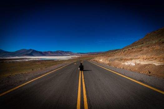 Lonely traveler sitting on a vast desert highway under a clear blue sky, with mountainous backdrop.