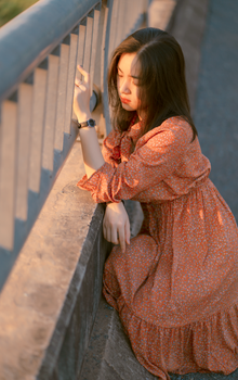 一位年轻女子穿着橙色连衣裙，在温暖的日光下，沉思地靠在栏杆上