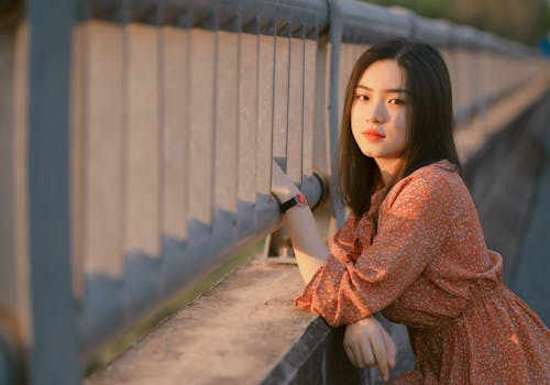 Woman in Orange Long Sleeve Dress Leaning on Concrete Fence With Railings