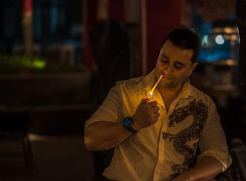 Free A man in a dragon-print shirt lights a cigarette in an outdoor café at night. Stock Photo