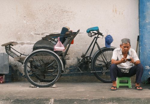 Free Elderly man sitting on a stool next to a bicycle rickshaw, using a phone. Stock Photo
