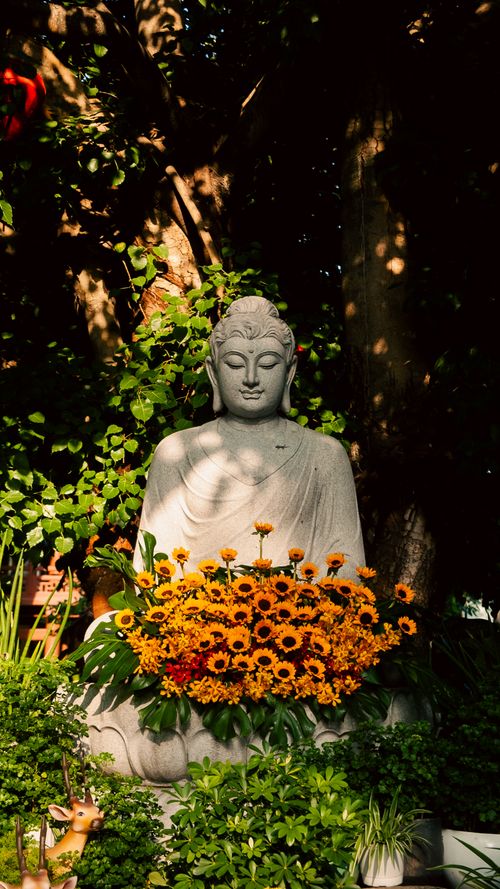 Free Buddha statue surrounded by greenery and vibrant flowers, creating a tranquil outdoor ambiance. Stock Photo