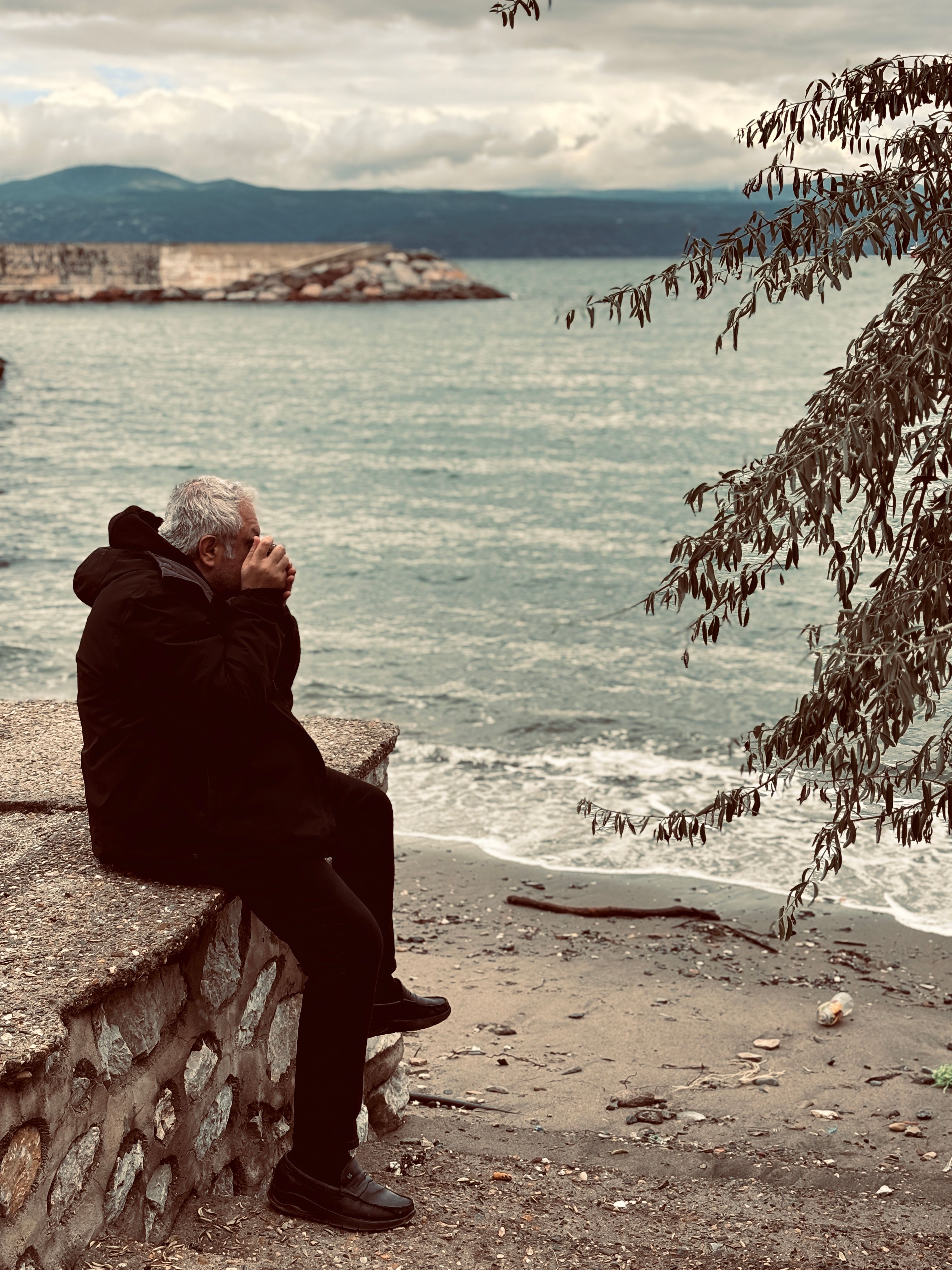 Older man sitting quietly by calm water, hands near face in thoughtful posture