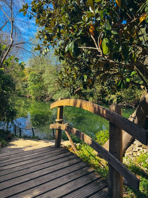 Free Peaceful wooden bridge over a calm river surrounded by lush spring greenery under a bright blue sky. Stock Photo