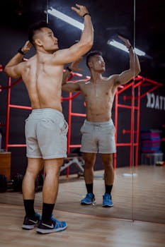 Muscular man in shorts poses confidently in front of gym mirror showcasing physique.