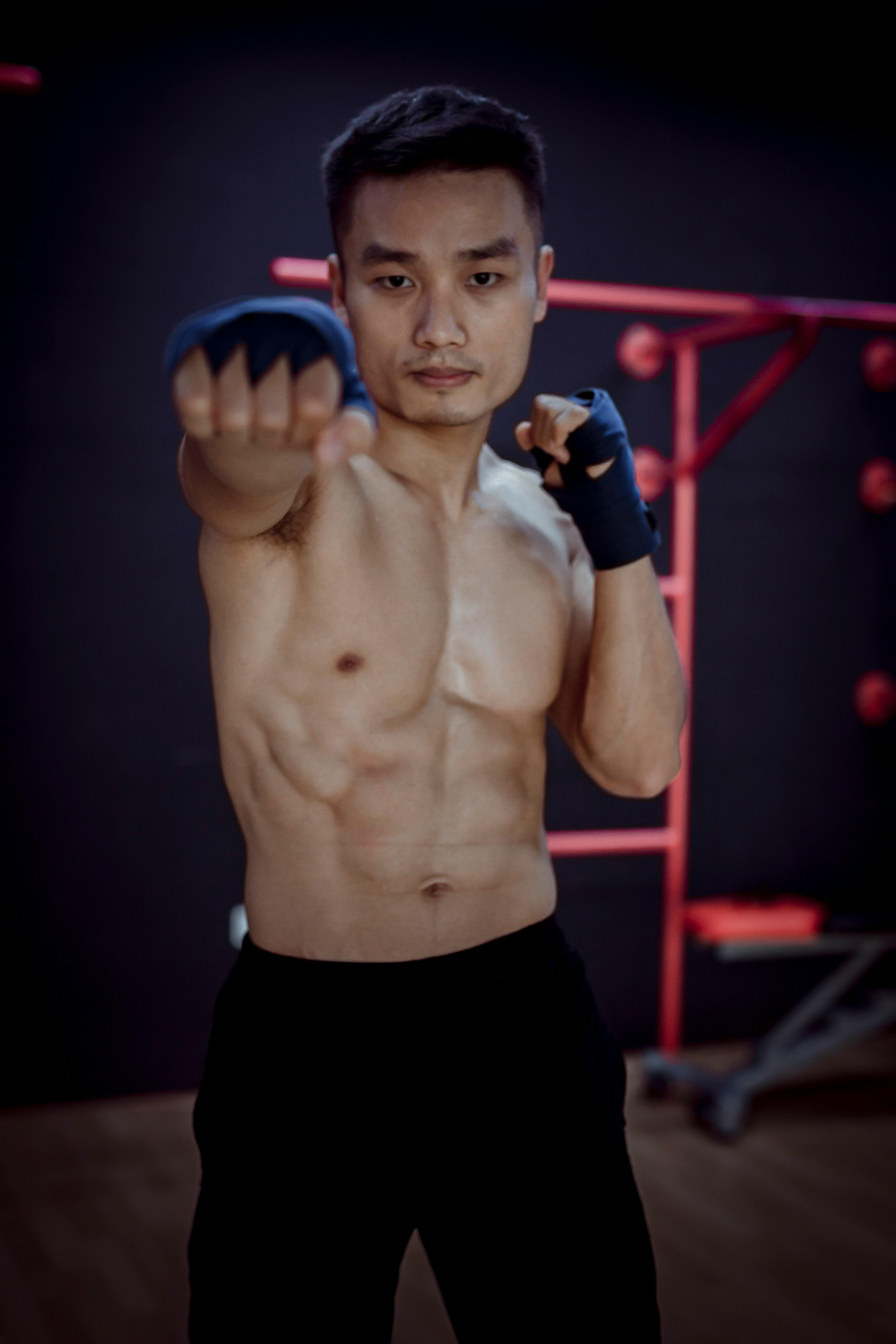 Serious young shirtless muscular male fighter in black pants and ace bandages confidently looking at camera while doing Thai boxing