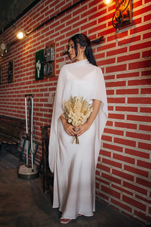 Free Woman in elegant white dress holds dried flowers against a brick wall in a stylish indoor setting. Stock Photo