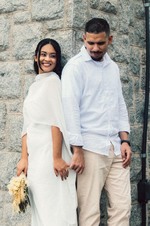Free A romantic couple in white clothing leaning against a textured stone wall, holding hands joyfully. Stock Photo