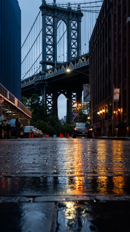 Free Dramatic dusk view of the Manhattan Bridge from a wet cobbled street in Brooklyn, reflecting city lights. Stock Photo