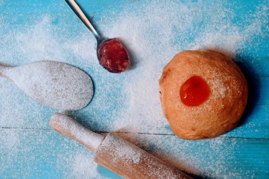 A freshly baked doughnut with jam and baking tools dusted with flour on a vibrant blue wooden surface.