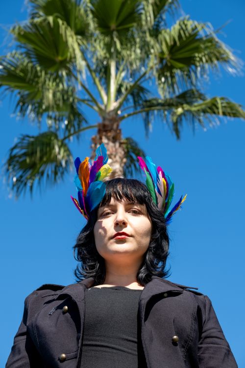 Free A woman wearing a colorful feather headpiece poses under a palm tree against a blue sky. Stock Photo