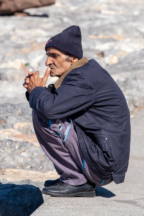 Free Elderly man crouching outdoors in thoughtful pose, wearing beanie and dark coat on stone pavement. Stock Photo