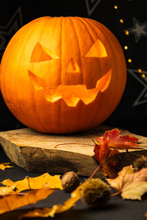 Free Carved pumpkin with a spooky face sitting on a wooden block, surrounded by autumn leaves. Stock Photo