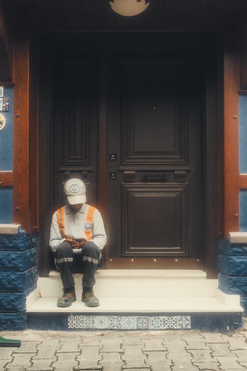 Free A solitary worker sits outside a detailed brown door, engrossed in their phone. Stock Photo