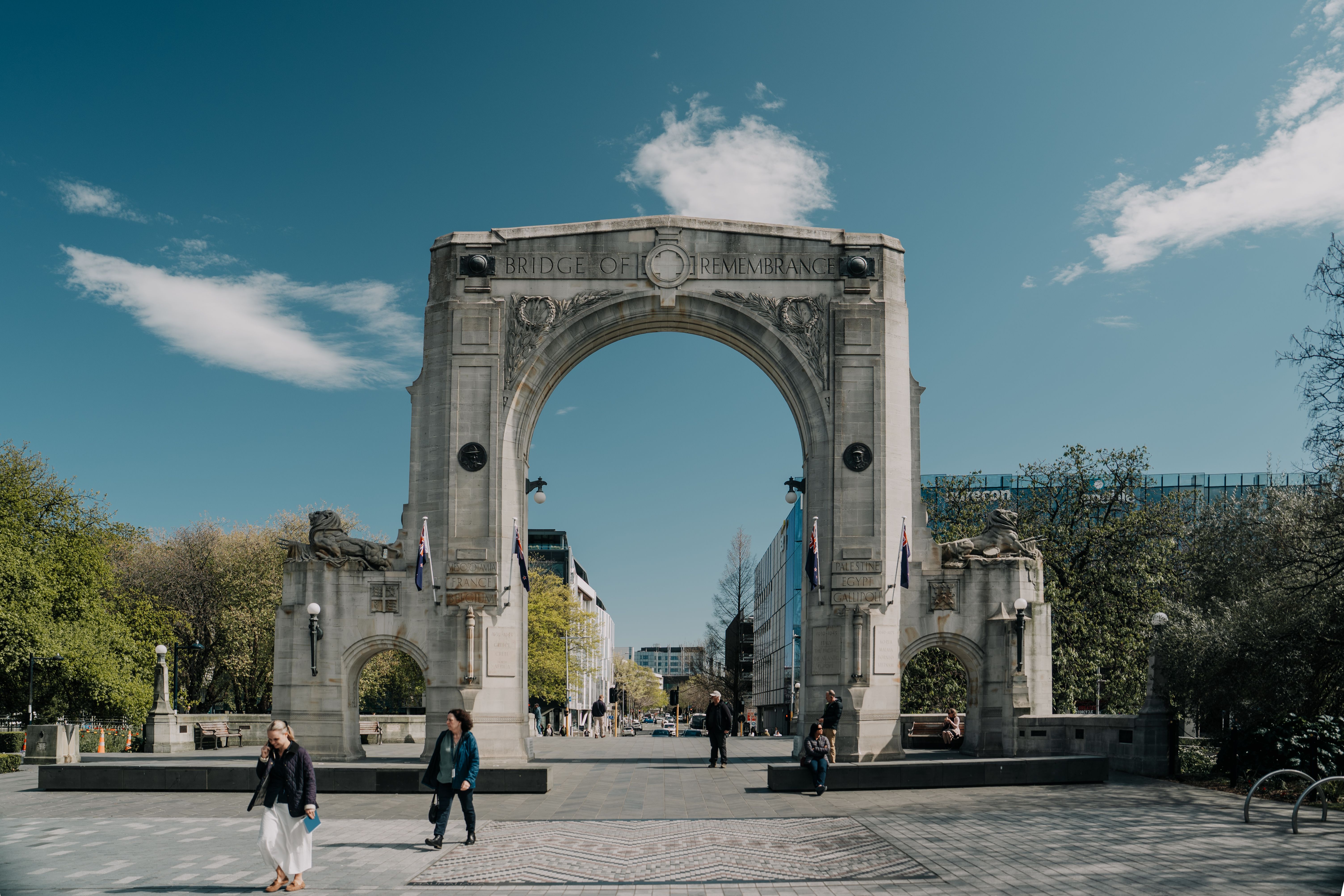 The historic Bridge of Remembrance in Christchurch on a sunny day, featuring pedestrians and lush greenery.
