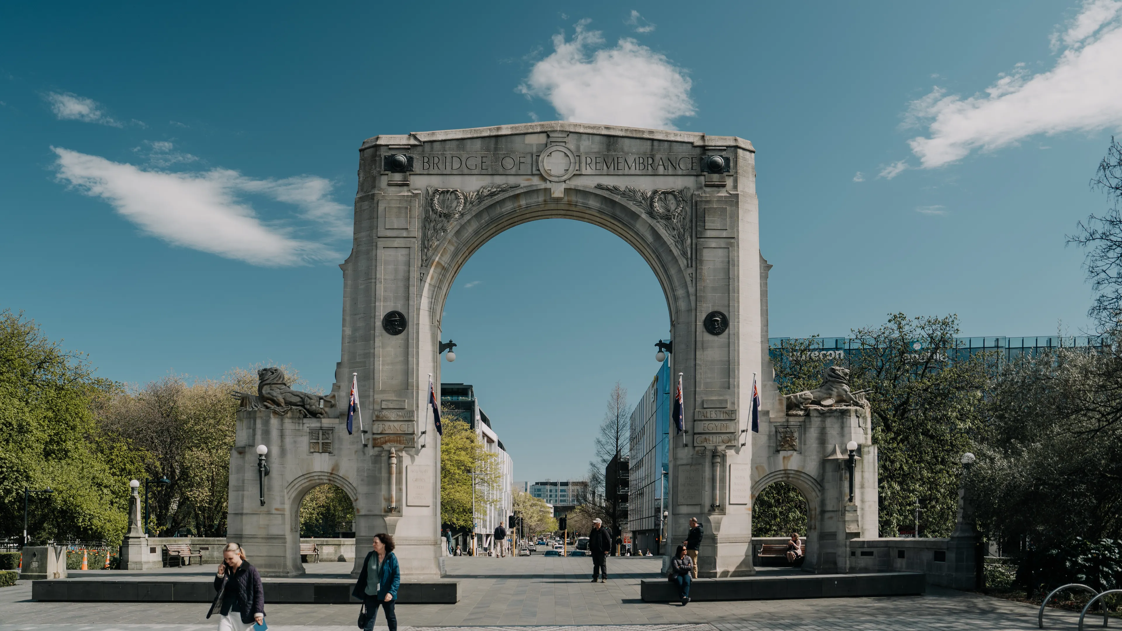 The historic Bridge of Remembrance in Christchurch on a sunny day, featuring pedestrians and lush greenery.