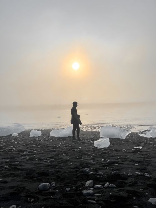 Free A person stands silhouetted by ice formations on a black sand beach during a foggy sunset in Iceland. Stock Photo