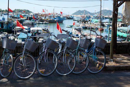 Free Rows of bicycles with baskets lined up by a vibrant harbor filled with fishing boats under sunny skies. Stock Photo