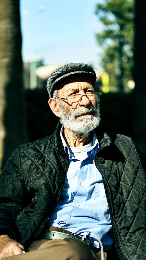 Free Portrait of an elderly man sitting outdoors in sunlight wearing a hat and glasses. Stock Photo