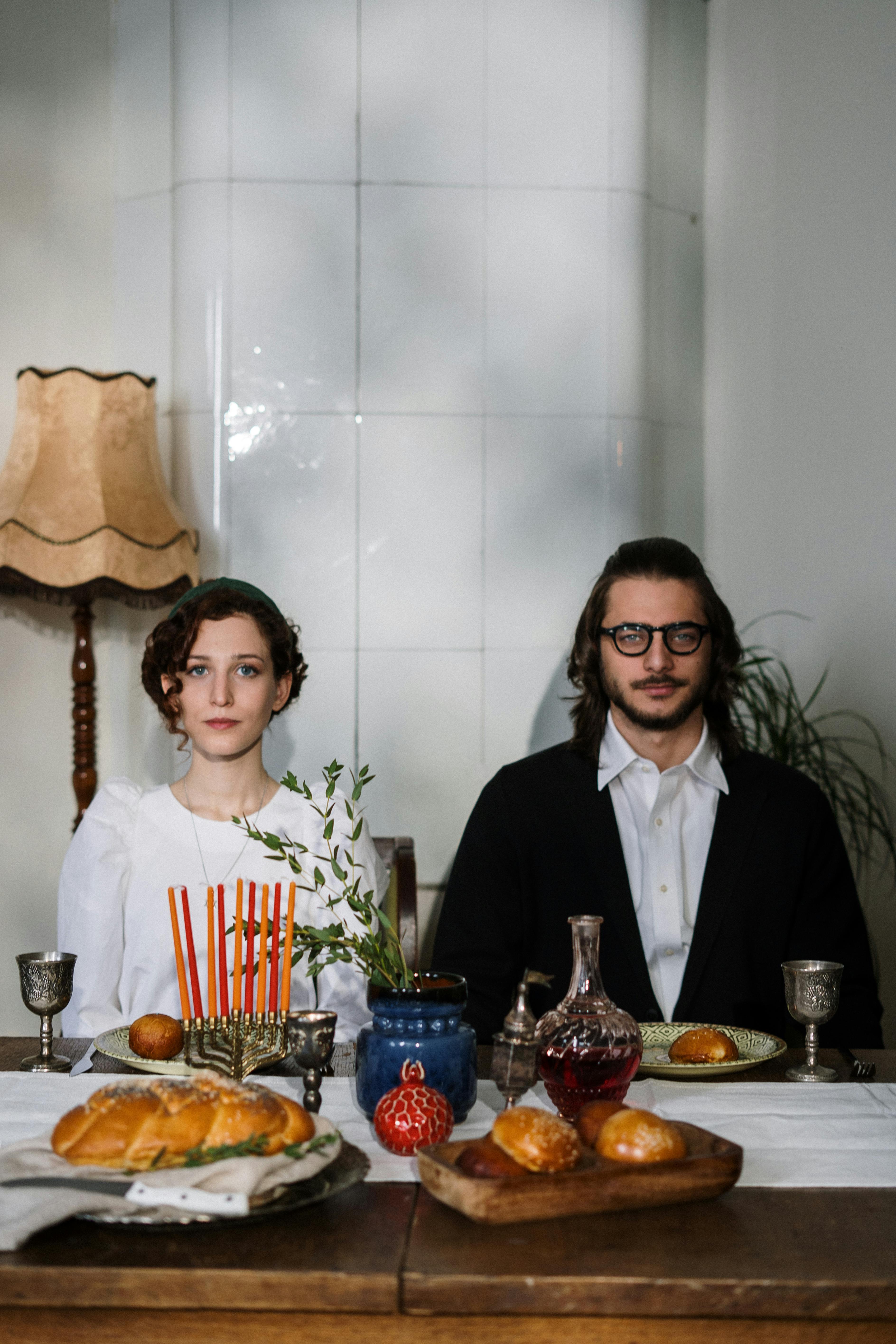 Orthodox Jewish couple at a Shabbat dinner table with traditional food and menorah in a cozy home setting.