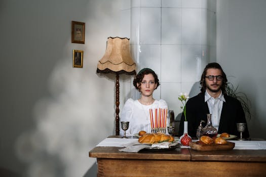 A couple in traditional attire at a table with challah and a menorah, creating a warm ambiance.