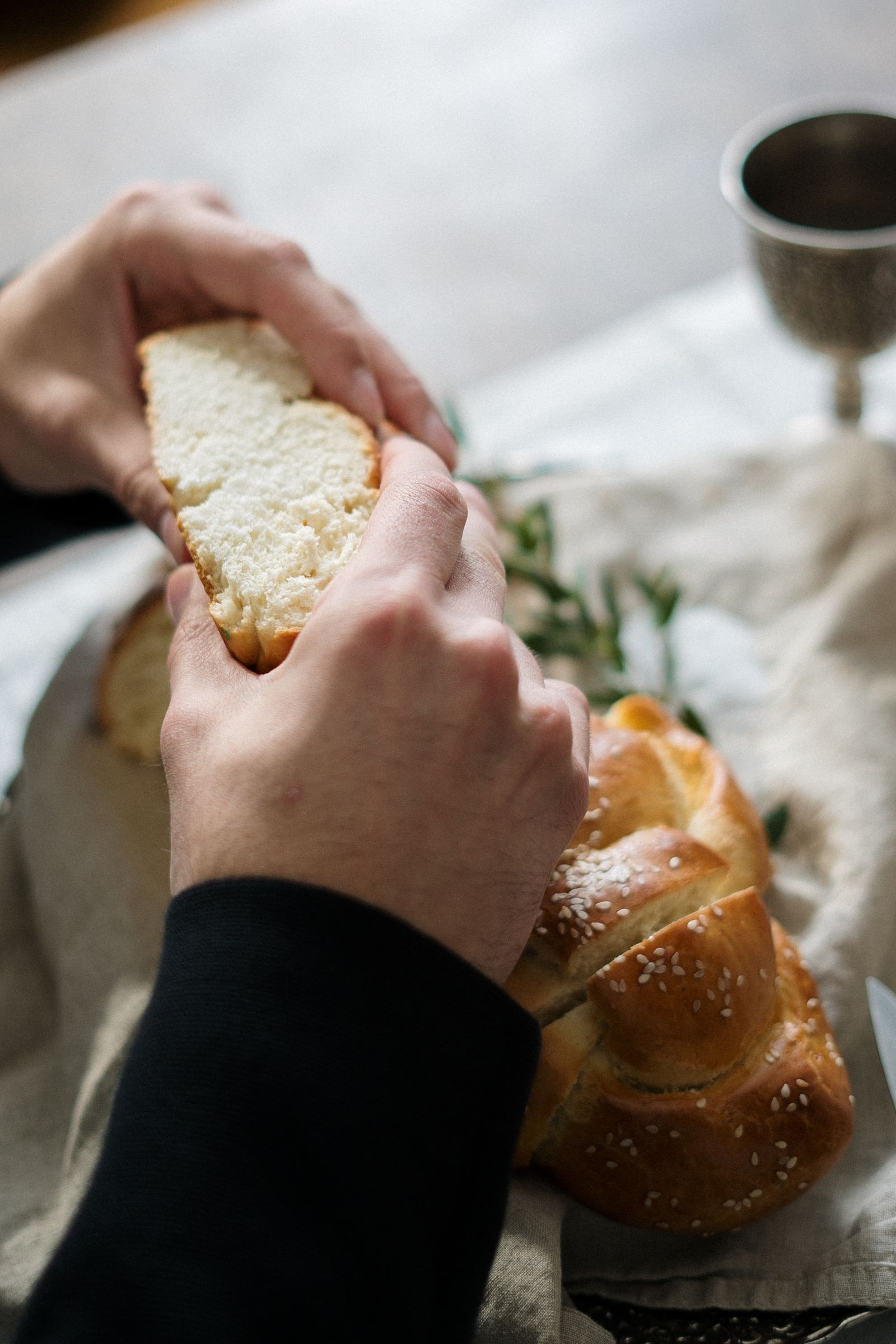 Hands tearing a piece from a braided challah next to a kiddush cup